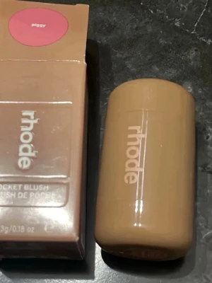 Rhode Pocket Blush Piggy - Rhode Pocket Blush i färgen Piggy är ett krämrouge i en smidig, beige cylinderformad förpackning. Färgen är en mjuk rosa ton som ger kinderna ett fräscht och naturligt glow. Perfekt att ha med i väskan för snabba touch-ups. Innehåller 5,3g.