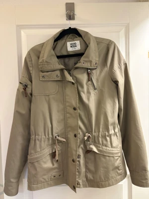 Beige vindjacka från Vero Moda - Stilren beige vindjacka från Vero Moda med dragsko i midjan, flera fickor med dragkedja och knappstängning framtill. Jackan har hög krage och detaljer i brunt läderimitation på dragkedjorna. Perfekt för blåsiga dagar och enkel att matcha.