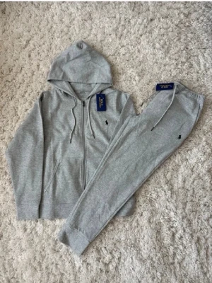 Grått loungewear-set från Polo Ralph Lauren - Snyggt och bekvämt loungewear-set i ljusgrått från Polo Ralph Lauren. Setet består av en hoodie med dragkedja och fickor samt matchande mjukisbyxor med dragsko i midjan. Båda plaggen har den klassiska Polo-loggan broderad på bröstet och benet.