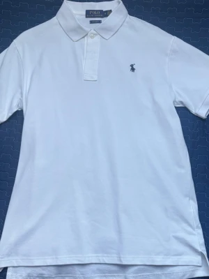 Vit pikétröja från Ralph Lauren - Klassisk vit pikétröja från Ralph Lauren med blå broderad logga på bröstet. Tröjan har korta ärmar, krage och två knappar vid halsen. Tillverkad i mjuk bomull som känns skön mot huden. Perfekt för en clean och stilren look.