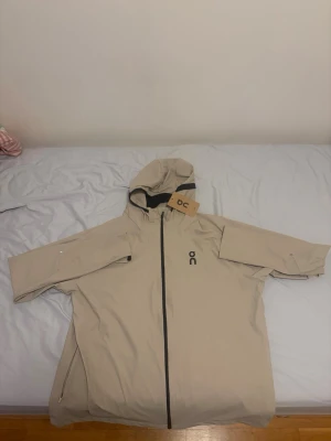 Beige regnjacka från On med huva - Snygg beige regnjacka från On med hel dragkedja framtill och justerbar huva. Jackan har svarta detaljer och logga på bröstet. Tillverkad i ett vattenavvisande material som passar perfekt för regniga dagar. Praktiska fickor med dragkedja på sidorna. STORLEKEN ÄR XXL MEN SKA PASSA EN I XL - jag har L och den är lite för stor
