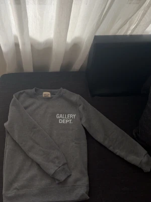 Grå sweatshirt från Gallery Dept. - Snygg grå sweatshirt från Gallery Dept. med vit logga tryckt på bröstet och stor text på ryggen. Klassisk rund halsringning och ribbade muddar vid ärmslut och nederkant. Perfekt för en avslappnad streetwear-look.