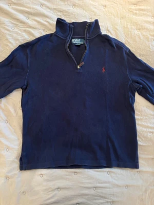 Marinblå halvzip från Polo Ralph Lauren - Klassisk marinblå tröja med halv dragkedja och hög krage från Polo Ralph Lauren. Tröjan har lång ärm och den ikoniska röda logotypen broderad på bröstet. Perfekt för lager på lager och en stilren look.