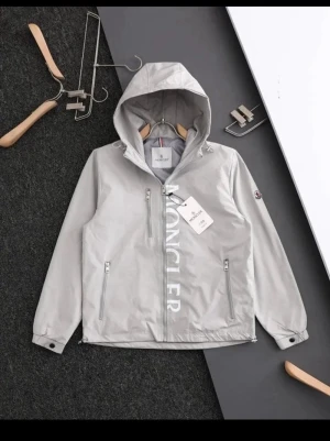 Grå vindjacka från Moncler med huva - Snygg grå vindjacka från Moncler med huva och dragkedja framtill. Jackan har stora vita Moncler-bokstäver längs dragkedjan, två sidofickor med dragkedja och en bröstficka. Klassisk Moncler-logga på ärmen och justerbara muddar.