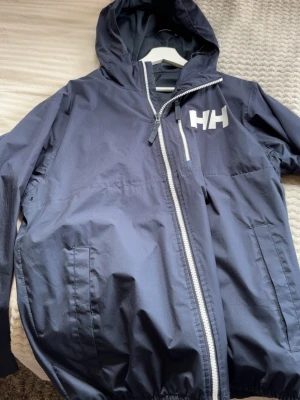 Mörkblå vindjacka från Helly Hansen - Snygg mörkblå vindjacka från Helly Hansen med vit HH-logga på bröstet. Jackan har huva, dragkedja framtill, två sidofickor och meshfoder för extra komfort. Perfekt för blåsiga dagar och enkel att matcha med olika outfits. Passar Medium också
