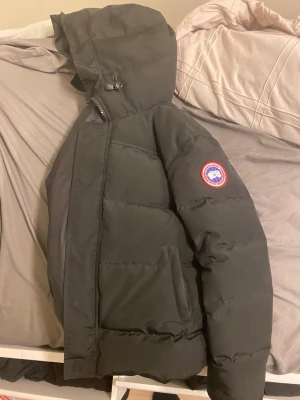 Svart pufferjacka från Canada Goose - Säljer en svart pufferjacka från Canada Goose med huva och klassisk logga på ärmen. Jackan har dragkedja och tryckknappar framtill samt två sidofickor. Perfekt för kalla vinterdagar och riktigt varm tack vare tjock vaddering.