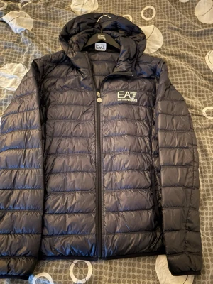 Svart pufferjacka från EA7 Armani - Snygg svart pufferjacka från EA7 Emporio Armani med huva och dragkedja framtill. Jackan har ett stilrent EA7-logotryck på bröstet och quiltad design som ger en modern look. Perfekt för kalla dagar och riktigt skön att bära.