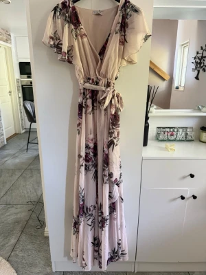 Ljusblommig långklänning med volangärm - Romantisk långklänning i ljus ton med blommigt mönster i rosa och lila. Klänningen har omlottmodell, v-ringning, volangärmar och ett knytband i midjan. Perfekt för sommarens alla tillfällen.