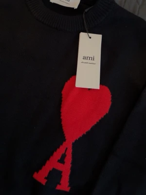 Ami Paris tröja - Snygg svart stickad tröja från Ami med röd hjärt- och A-detalj på bröstet. Tröjan har rund halsringning, ribbade muddar och nederkant. Perfekt för dig som gillar stilrena plagg med en twist.