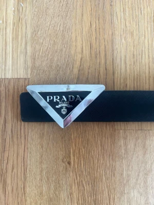 Svart skärp med triangel spänne - Snyggt svart skärp med en stor, trekantig silverfärgad logga framtill. Skärpet är gjort i skinn och har en stilren design som verkligen sticker ut. Perfekt accessoar för att lyfta vilken outfit som helst. 