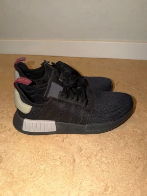 Svarta Adidas NMD_R1 sneakers - Snygga svarta Adidas NMD_R1 sneakers med stickad ovandel, svarta och grå detaljer samt vit och grå sula. Skorna har röda detaljer på hälen och klassiska tre ränder på sidan. Perfekta för en streetwear-look och riktigt bekväma med sin flexibla design.