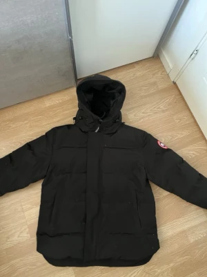 Svart pufferjacka från Canada Goose - Säljer en svart pufferjacka från Canada Goose med huva och klassisk logga på ärmen. Jackan har dragkedja och vindslå framtill samt stora fickor. Perfekt för kalla vinterdagar och riktigt varm tack vare dunfyllning. Snygg och stilren design som passar till det mesta.
