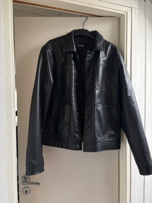 Svart skinnjacka från Pull&Bear - Stilren svart skinnjacka från Pull&Bear med klassisk krage och raka linjer. Jackan har två stora fickor framtill och stängs med knappar. Perfekt för dig som vill ha en tidlös och cool look. (ej äkta läder). Köpt i Barcelona för 600kr