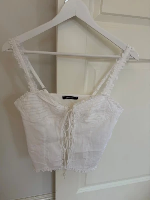 Vit snörad korsettopp från Gina Tricot - Säljer en söt vit korsettopp från Gina Tricot med snörning framtill och spetsdetaljer längs axelbanden och nederkanten. Toppen är ärmlös och har en croppad passform som ger en trendig look.