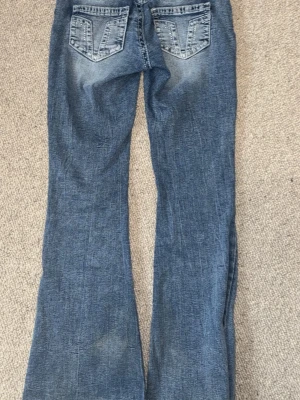 Blå bootcut jeans med slitningar - Ett par blå bootcut jeans med snygga slitningar och tvättade detaljer. Jeansen har klassiska bakfickor med V-formade sömmar och lätt utsvängda ben. Perfekta för en avslappnad och trendig look.