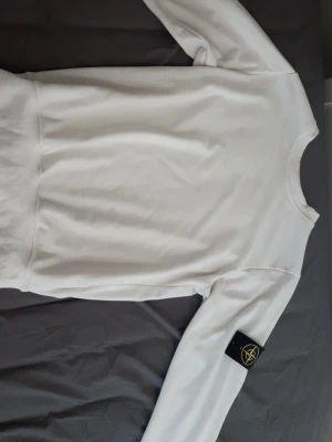 Vit sweatshirt från Stone Island - Säljer en clean vit sweatshirt från Stone Island med den klassiska svarta patchen med gul och grön kompass på vänster ärm. Tröjan har rund halsringning, ribbade muddar och är tillverkad i mjukt bomullsmaterial. Perfekt för dig som gillar streetwear och snygga detaljer.