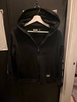 EA7 hoodie - Svart hoodie från EA7 Emporio Armani med dragkedja framtill och huva. Ärmarna har vita logotexter längs sidorna och en liten EA7-logga finns på magen. Perfekt för en sportig och stilren look. Storlek 160cm men passar mig som är 169cm. Skulle dock säga att den passar bäst om man är 165 typ.
