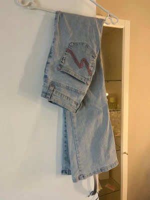 Ljusblå jeans från Nelly - Säljer ett par ljusblå jeans från Nelly med raka ben och cool fickdetalj med röd zigzag-söm. Klassisk femficksmodell i mjukt denimtyg och dragkedja framtill. Perfekta för en avslappnad och trendig look.