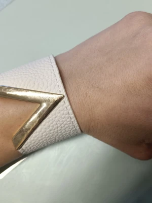 Brett vitt armband med gulddetalj - Stilrent och brett armband i vitt läder med en snygg V-formad gulddetalj framtill. Armbandet har struktur i materialet och stängs med tryckknappar i guld på sidan. Perfekt accessoar för att lyfta din outfit.