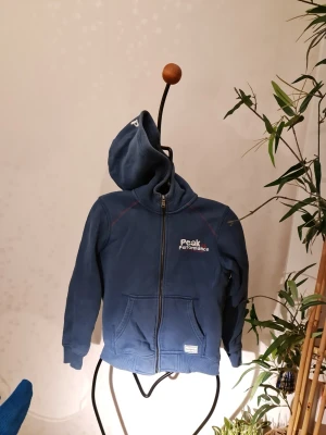 Blå hoodie från Peak Performance - Säljer en blå hoodie från Peak Performance med dragkedja framtill och broderad logga på bröstet. Hoodien har två fickor fram och en klassisk huva. Perfekt för dig som gillar sportig och avslappnad stil.