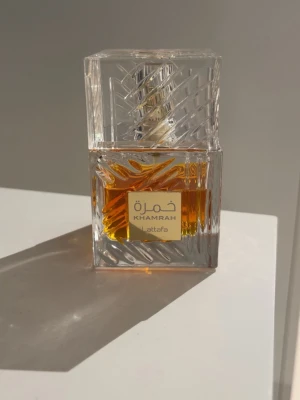 Khamrah parfym från Lattafa - Khamrah parfym från Lattafa i en elegant, fyrkantig glasflaska med mönstrad design. Flaskan är genomskinlig och visar den bärnstensfärgade vätskan inuti. Ungefär 70% kvar