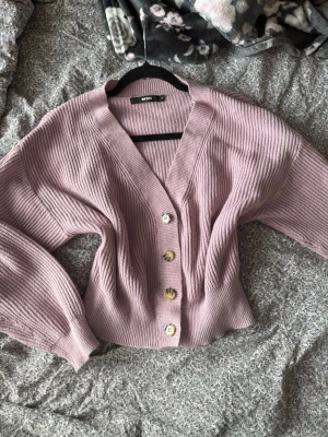 Ljuslila kofta från Bik Bok - Säljer en ribbstickad ljuslila kofta från Bik Bok med v-ringning och fyra stora beige knappar framtill. Koftan har ballongärmar och är något croppad i modellen. Perfekt att slänga över en topp för en chill look.