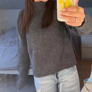 Mörkgrå stickad tröja med polokrage - Mysig mörkgrå stickad tröja med hög polokrage och långa ärmar. Tröjan har en rak passform och ribbstickade muddar vid ärmslut och nederkant. Perfekt för lager-på-lager och enkel att matcha med jeans.