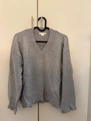 Grå stickad tröja från Gina Tricot - Snygg grå stickad tröja från Gina Tricot med djup v-ringning och breda ribbade muddar vid ärmslut och nederkant. Perfekt för lager på lager och enkel att matcha med olika outfits. Mjuk och bekväm kvalitet.