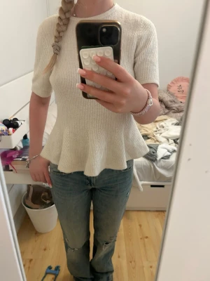 Vit ribbad topp med volangkant - Säljer en söt vit ribbad topp med volangkant nedtill. Toppen har korta ärmar och rund halsringning. Materialet är stickat och ger en mjuk och bekväm känsla. Storlek M men passar mig som brukar ha xs eller s