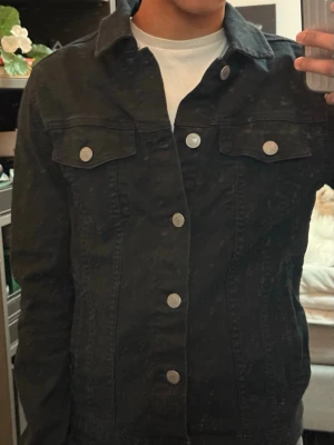 Svart jeansjacka från Jack & Jones - Snygg svart jeansjacka i storlek XS från Jack & Jones med klassisk krage, silverfärgade knappar och två bröstfickor med lock. Jackan har en rak passform och är tillverkad i slitstarkt jeansmaterial. Perfekt för en cool och avslappnad stil.