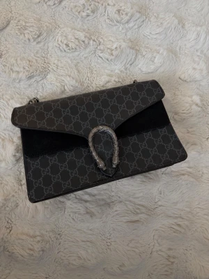 Svart axelväska  - Snygg svart axelväska med klassiskt GG-mönster och silverfärgad kedja. Väskan har en unik hästskospänne framtill och är tillverkad i canvas med detaljer i mocka. Perfekt för dig som vill ha en trendig och lyxig accessoar.