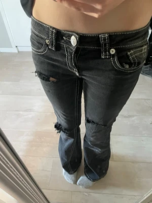 Svarta bootcut jeans med slitningar - Säljer ett par svarta bootcut jeans med vita kontrastsömmar och slitna detaljer vid knä och lår. Jeansen har fem fickor och dekorativa sömmar på bakfickorna. Perfekta för en avslappnad och trendig look.