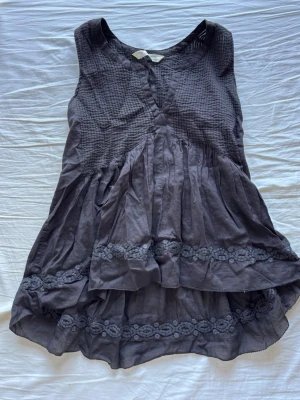 Mörkgrå ärmlös blus med spetsdetaljer - Säljer en flowy mörkgrå ärmlös blus med vackra spetsdetaljer och volanger. Toppen har en lätt rynkad struktur upptill och dubbla lager tyg nedtill. Storlek 0 som motsvarar xs
