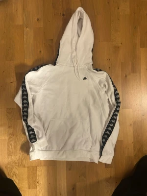Vit hoodie från Kappa med logoband - Snygg vit hoodie från Kappa med klassiska svarta logoband längs ärmarna och stor huva. Hoodien har en stor magficka och Kappa-logga broderad på bröstet. Tillverkad i mjukt bomullsmaterial som är skönt mot huden.