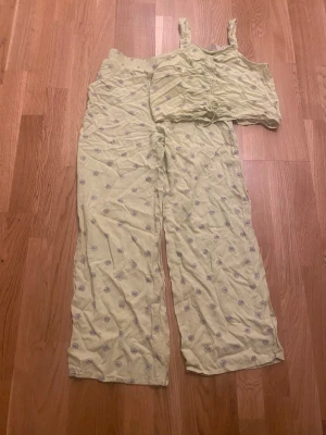 Gult blommigt set med topp och byxor - Säljer ett gulligt set med matchande topp och byxor i ljusgul färg med små vita och lila blommor. Toppen har breda axelband och rynkdetaljer framtill, byxorna är vida och har resår i midjan. Perfekt för en avslappnad och söt look.