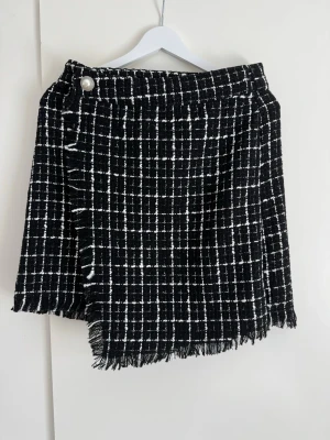 Svart vit mini kjol, storlek 38 - Svart och vit rutig kjol med fransiga kanter. Kjolen har en omlottdesign framtill som skapar en asymmetrisk nederkant. En dekorativ pärlemorknapp med silverdetaljer pryder ena sidan av midjan. Dragkedja baktill för smidig stängning.  Length: 48 cm  Buy shipping label or pick it up from Farsta Centrum in Stockholm.