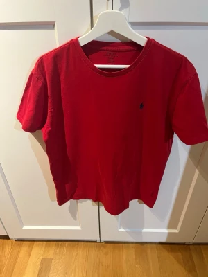 Röd t-shirt från Polo Ralph Lauren - Röd t-shirt från Polo Ralph Lauren med klassisk passform och liten broderad logga på bröstet. Tillverkad i mjuk bomull och har rund halsringning samt korta ärmar. Perfekt för en avslappnad och stilren look.