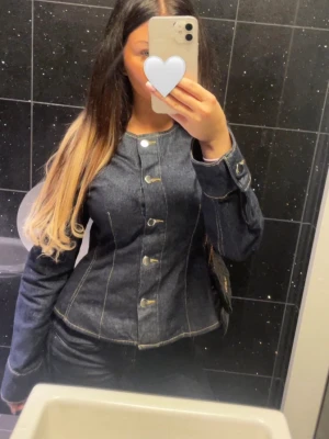 Mörkblå figursydd jeansjack/tröja - Snygg mörkblå jeansjacka/tröja med figursydd passform och dekorativa sömmar. Jackan har knappar framtill och långa ärmar. Perfekt för dig som vill ha en trendig och stilren look.