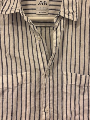Randig kortärmad skjorta från Zara - Snygg vit skjorta från Zara med blåa ränder. Skjortan har korta ärmar, två bröstfickor och knäppning framtill. Relaxed fit och tillverkad i ett lätt och luftigt linnematerial – perfekt för varma dagar.
