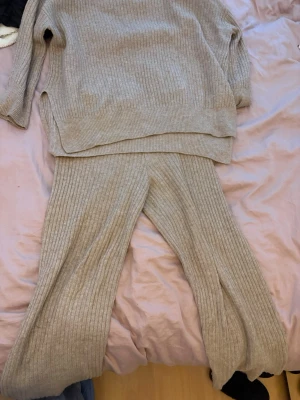 Beige ribbat loungewear-set från H&M - Mysigt loungewear-set från H&M i beige ribbstickat material. Setet består av en långärmad tröja med slits i sidorna och matchande vida byxor. Perfekt för chill dagar hemma. Materialet är mjukt och stretchigt för extra komfort.