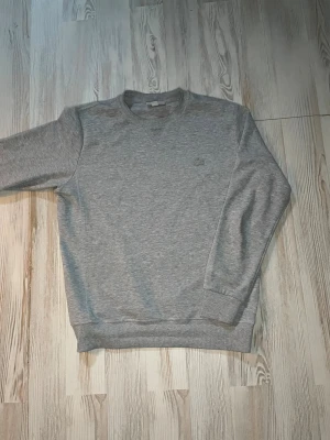 Grå sweatshirt från Lacoste - Klassisk grå sweatshirt från Lacoste med rund halsringning och diskret krokodillogga broderad på bröstet. Tröjan har ribbade muddar vid ärmslut och nederkant. Perfekt för en avslappnad och stilren look.