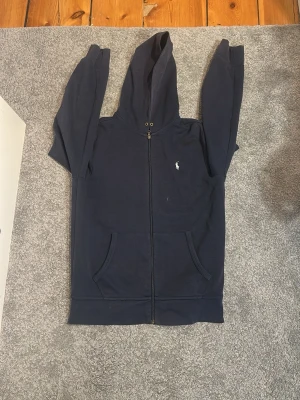 Mörkblå hoodie från Polo Ralph Lauren - Mörkblå hoodie med dragkedja från Polo Ralph Lauren. Klassisk design med huva, två fickor framtill och den ikoniska vita logotypen broderad på bröstet. Perfekt för en avslappnad och stilren look.