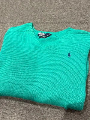 Turkos stickad tröja Polo Ralph Lauren - Turkos stickad tröja från Polo Ralph Lauren med V-ringning och klassisk blå logga broderad på bröstet. Tröjan har långa ärmar och ribbstickade muddar vid ärmslut och nederkant. Perfekt för en stilren och avslappnad look. Finns några enstaka små fläkar därav priset.
