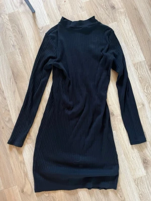 Svart ribbad långärmad klänning - En svart, ribbad klänning med lång ärm och hög hals. Klänningen har en tight passform och är tillverkad i ett stretchigt material som sitter snyggt på kroppen. Perfekt för dig som gillar en stilren och enkel look.