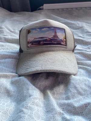 Beige truckerkeps med Paris-motiv - Säljer en beige truckerkeps med mesh på sidorna och ett coolt tryck framtill som visar en bil och Eiffeltornet i Paris. Kepsen har böjd skärm och justerbar baksida. Perfekt för dig som gillar streetwear och unika accessoarer.