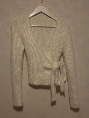 Vit fluffig omlottkofta - Supermjuk och fluffig vit stickad omlottkofta från SHEIN. Koftan har långa ärmar, knytband i sidan och en croppad passform. Materialet är en mix av polyamid och polyester som ger en mysig känsla och trendig look. Knappt använd🤍