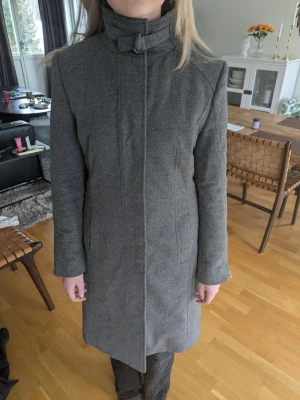 Grå ullkappa från Hugo Boss - Klassisk och tidlös grå kappa/rock i 90% ull, 10% kashmir med hög krage och spänne i halsen. Rak passform, diskreta fickor och stilren design. Perfekt för kyliga dagar när du vill hålla stilen enkel men trendig.