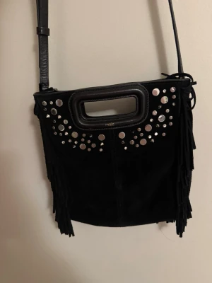 Svart axelremsväska med nitar från Maje - Cool svart handväska i mocka från Maje med silverfärgade nitar framtill, fransar på sidorna och både axelrem och handtag. Väskan har en rektangulär form och detaljer i läder. Jag har använt den två gången så nästan helt oanvänd.