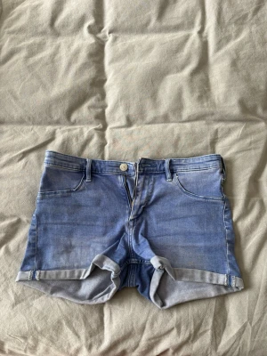  lågmidjade jeans shorts - Säljer ett par blå lågmidjade shorts från Bik Bok med klassisk femficksdesign och uppvikta benslut. Perfekta för varma dagar och enkel att matcha med olika toppar.