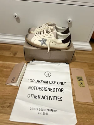 Golden Goose vita sneakers med glitter - Snygga vita sneakers från Golden Goose med silverglittrig stjärna på sidan och mörkbrunt glitter på hälen. Skorna har beige sula, vita skosnören och detaljer i mocka vid tån. Perfekta för dig som vill sticka ut med en unik och trendig look. Passar för 45.Har aldrig använt.